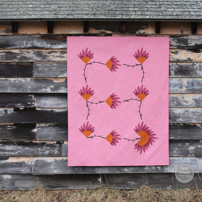 Daisy Chain Variation quilt hanging on a building with weather worn siding. Quilt is pink with foundation paper pieced sunflower blocks and dark thorny stems.