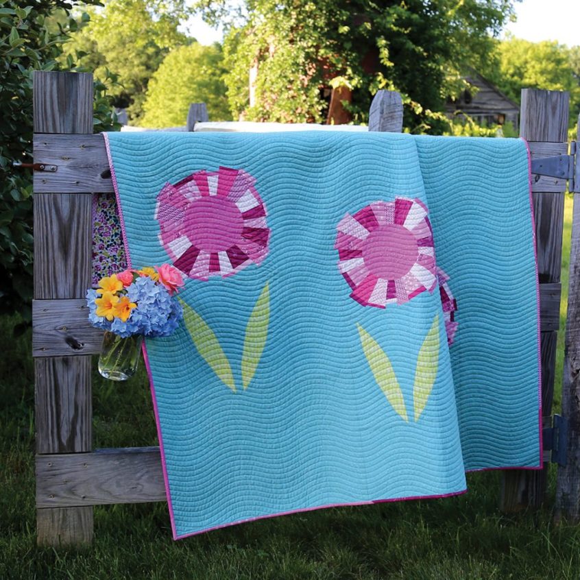 Mod Blooms Quilt hanging over wooden gate. Flowers are made from a scrappy assortment of berry colored prints.