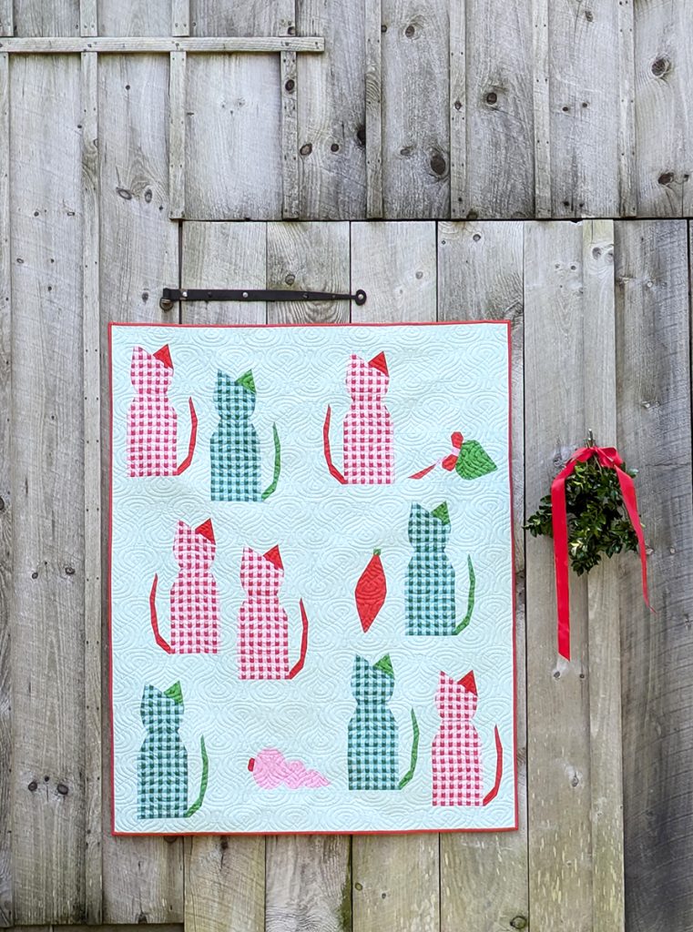 Christmas themed quilt with cats and vintage ornaments hanging on a rustic wood barn with a bunch of holly.