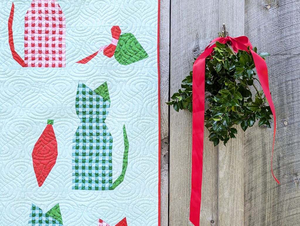 Christmas themed quilt with cats and vintage ornaments hanging on a rustic wood barn with a bunch of holly.