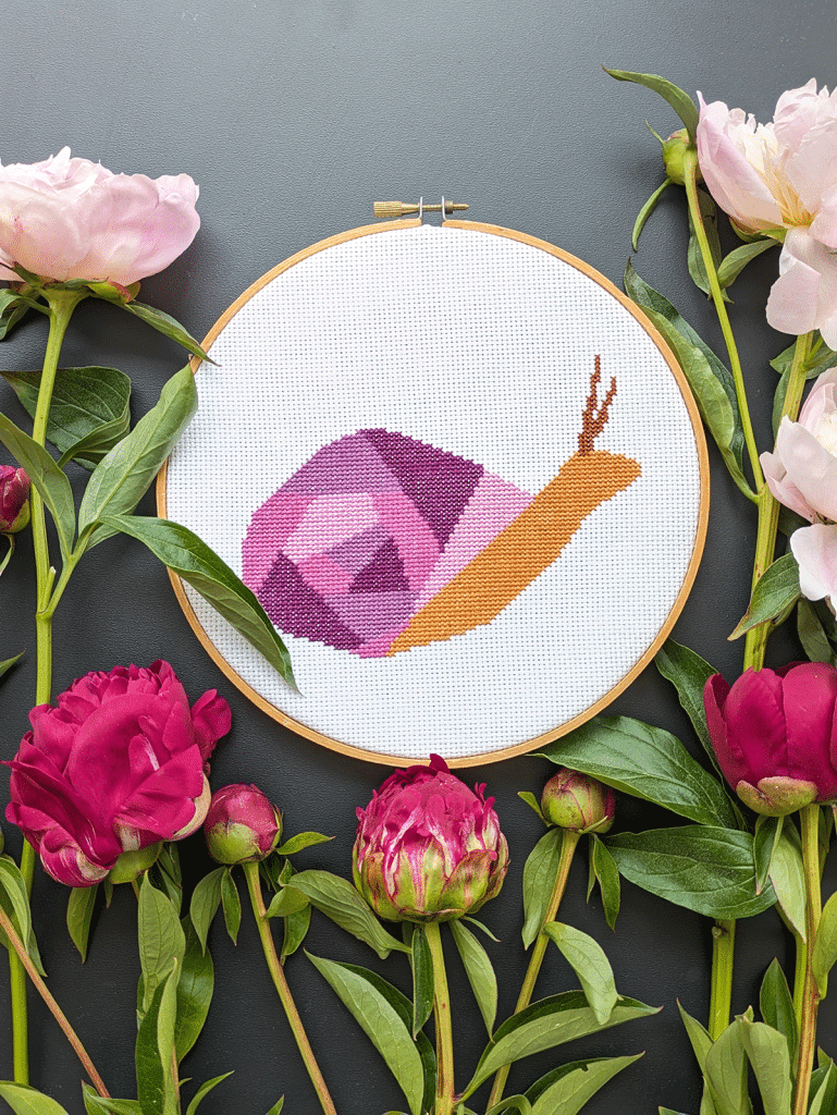 Garden snail cross stitch pattern in an embroidery hoop surrounded by peonies.
