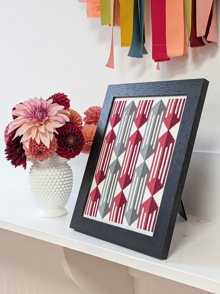 Caged cross stitch art in a black frame next to dahlias in a white vase