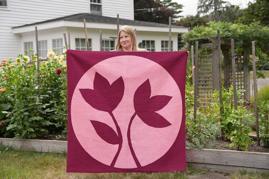 Amy holding her Flower Moon Quilt in front of her garden.